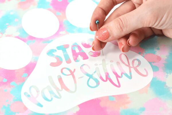 Learn the hack to get bubbles out of vinyl. A woman's hand hold a pin above the vinyl tie-dyed paw print that says "Stay Paw-sitive."