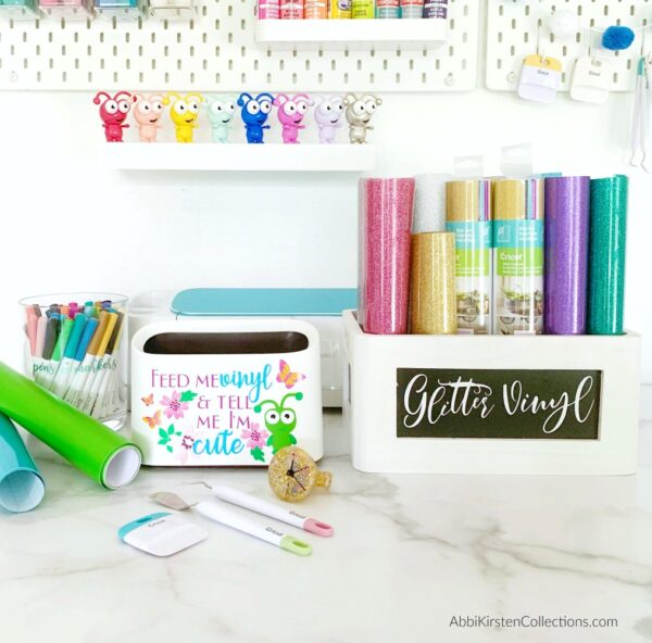 A closeup of a tabletop hosting several craft supplies like the vinyl scraps trashcan, markers and a wooden bin storing glitter vinyl rolls. Storing and organizing vinyl rolls is one tip to make vinyl and Cricut crafting easier. 