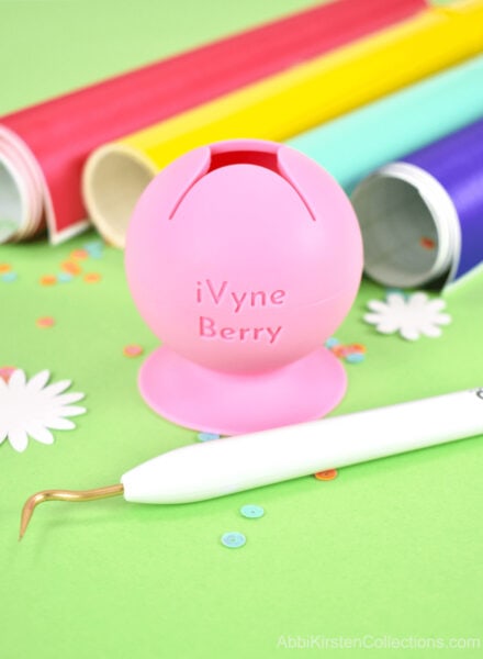 Tips for weeding Cricut vinyl include using this iVyne Berry Tool. A closeup of the pink berry tool, a standard weeding tool an primary color vinyl rolls on a green table top. 