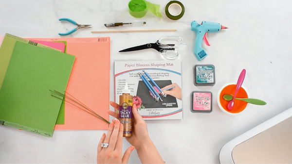 All the supplies needed to make this paper rose bouquet - paper, glue, curling dowel, and stem wire - sit on a white crafting workspace.