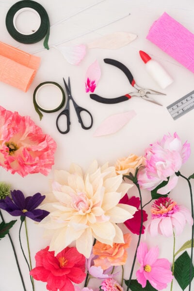 A flat-lay image of various crepe paper flowers in different shades of white, red, pink, and blue. Supplies to make the flowers lay scattered on a white background - scissors, wire cutters, clue, a ruler, and rolls of crepe paper.