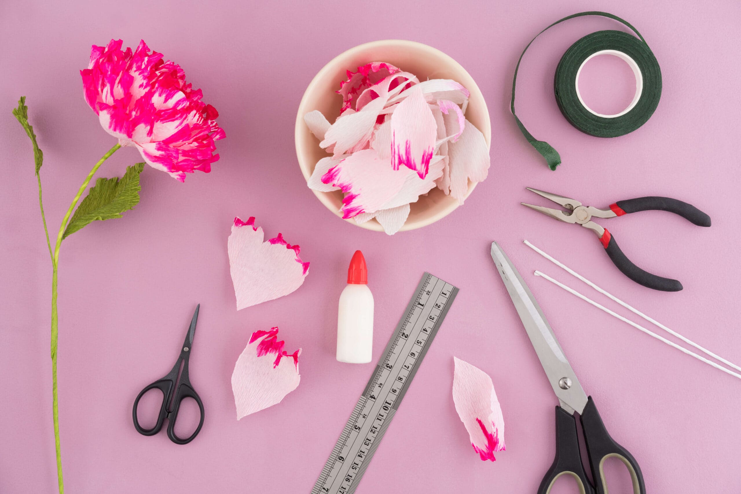 A flatlay of different supplies needed to make crepe paper flowers. White flower petals with pink-dyed tips fill a cream colored bowl. There's floral tape, wires, pliers, glue, a ruler, and scissors.