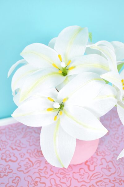 Bunches of handmade white crepe paper lilies with white and yellow flower centers fill a pink vase.