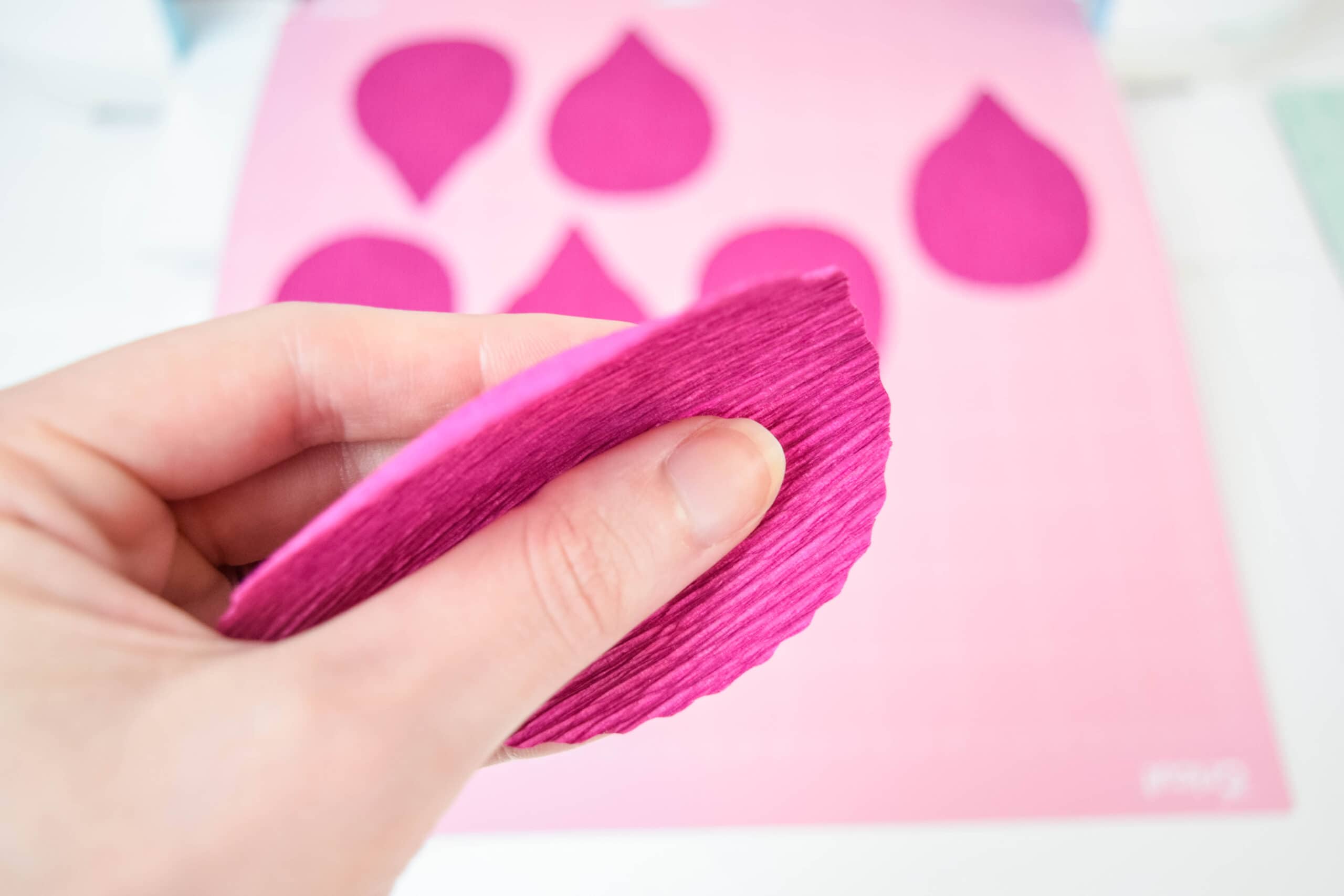 A woman rolls pink crepe paper flower petals between her thumb and pointer finger, gently curling and stretching the flower petal.