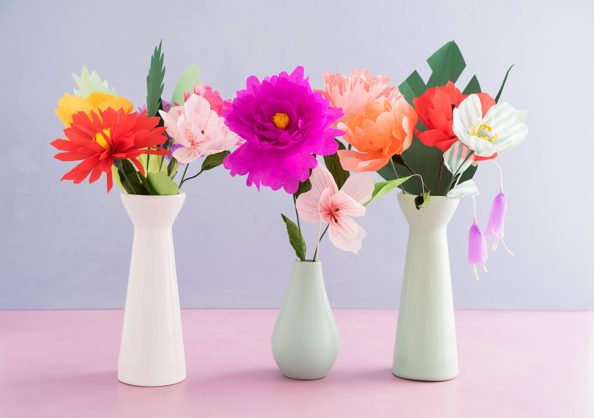 Three flower vases filled with colorful crepe paper flowers, sitting on a pink tabletop in front of a lavender background.