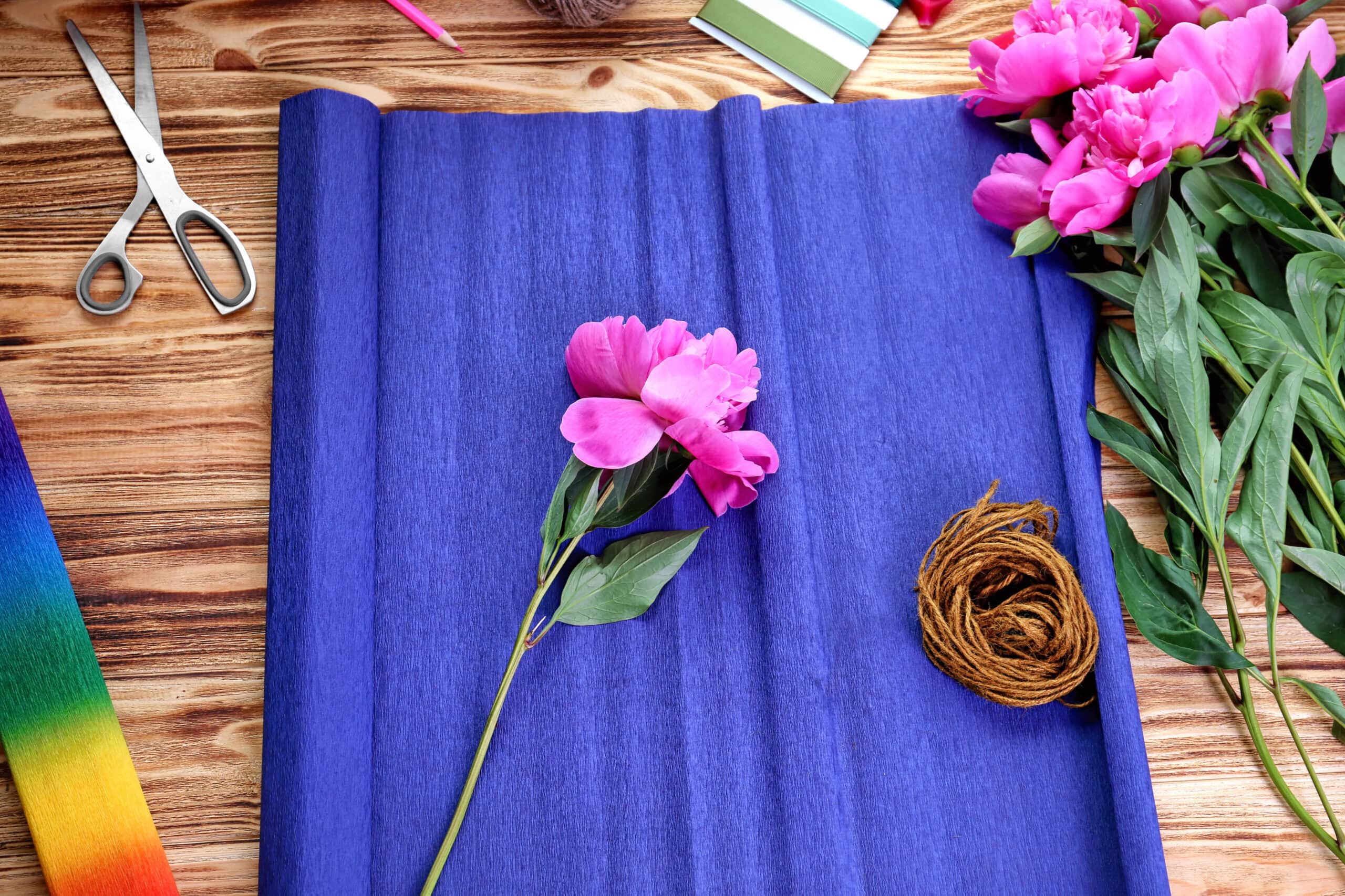 Purple crepe paper peonies sit on a spread-out roll of blue crepe paper. Also in the image are a pair of scissors, a bunch of twine, and a roll of rainbow crepe paper. The image is high saturated, so the colors are both and bright.