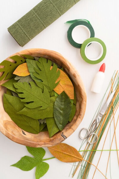 A bowl filled with green and gold crepe paper leaves, floral wire, scissors, floral tape, and a roll of green crepe paper.