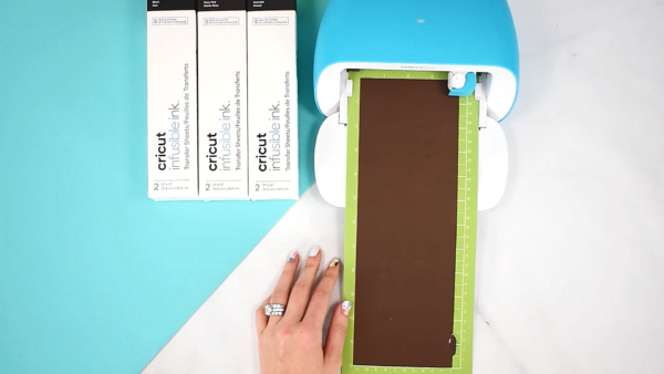 Abbi Kirsten's hand guides a sheet of brown Cricut Infusible Ink into a Cricut Joy. Three boxes of Cricut Infusible Ink are lined up next to the cutting machine. 