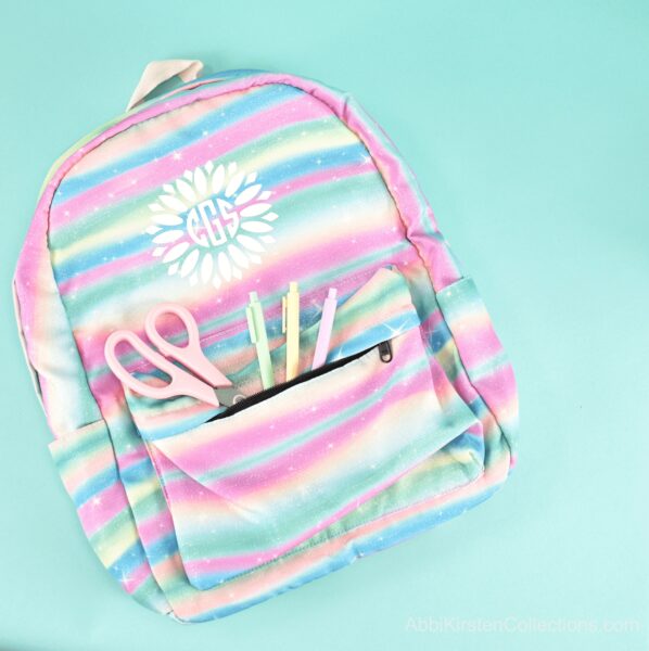 A pastel rainbow kids backpack with an iron-on vinyl decal on the front, packed with pens and scissors for back-to-school.