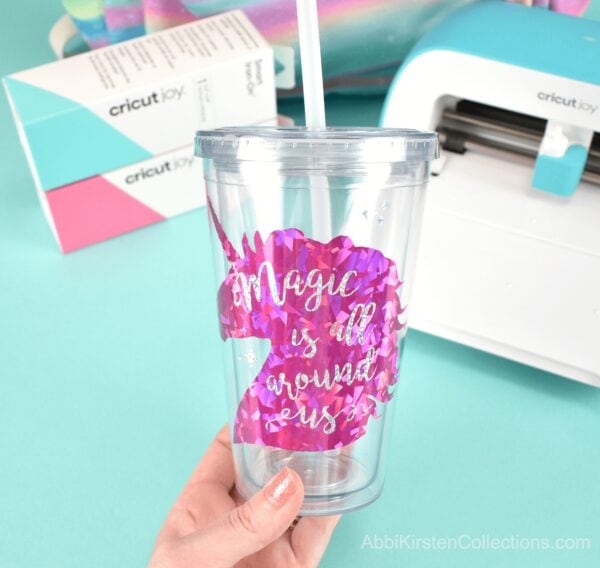 A clear acrylic cup with a holographic pink unicorn vinyl applied to the front. The cup is held up in front of a Cricut Joy cutting machine and boxes of Smart Iron-On vinyl.