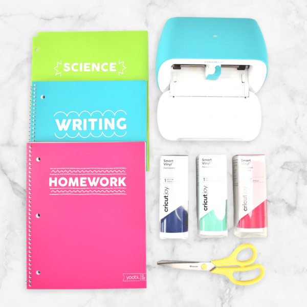 Three school notebooks lined up neatly next to a Cricut Joy cutting machine, rolls of Smart Vinyl, and scissors. The notebooks are lime green with a decal that says "vinyl", bright blue with a "writing" decal, and bright pink with a "homework" decal.