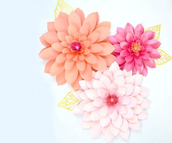 Three giant paper dahlia flowers made with light pink, orange, and dark pink paper. Each dahlia flower has a poppy center and yellow paper leaves.