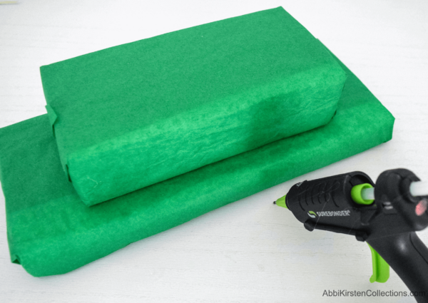 Two green tissue paper wrapped floral foam blocks stacked on top of each other, and a hot glue gun to glue them together.