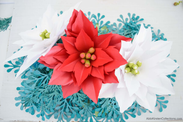 Three paper poinsettia flowers, two white and one red, placed in the floral foam blocks, surrounded by paper mistletoe leaves.