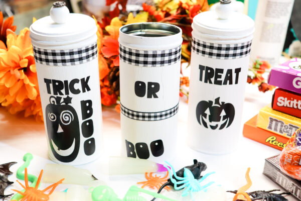 White and black tall cans decorated with black and white plaid, jack-o-lanterns and the words "Trick or treat" and "boo." These trick or treat cans are on a table with orange flowers, candy boxes and fake spiders. 