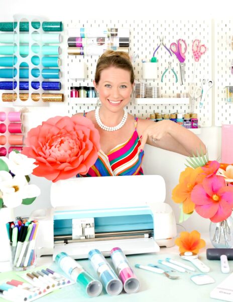 Abbi Kirsten smiles and points to the Cricut machine in front of her while in her craft room, surrounded by art supplies and paper flowers. 