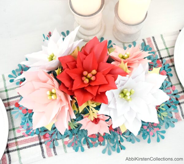 The completed paper poinsettia flower arrangement has four large paper poinsettias, two white, one red, and one pink, as well as two smaller pink poinsettia flowers. The flowers are surrounded by mistletoe and berry springs made from blue and red paper.