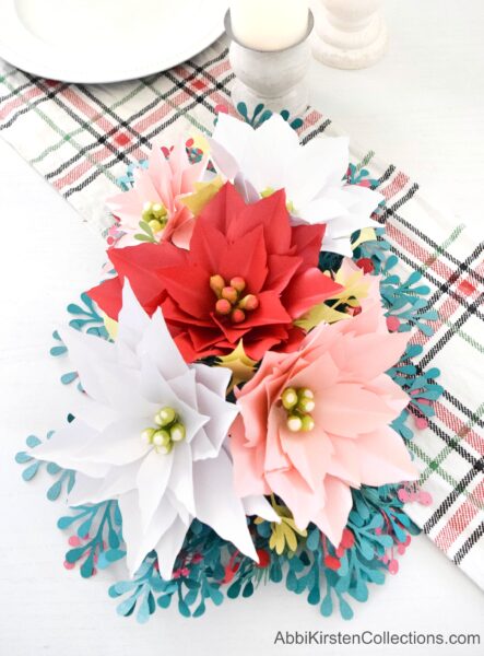 A paper poinsettia flower arrangement sits on a white table next to a plaid fabric table runner.