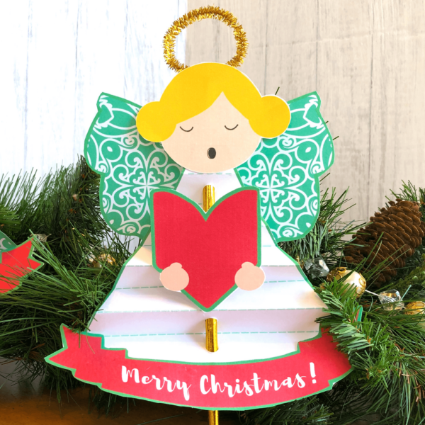 A blonde choral angel made with paper and paper straws sits against an evergreen garland. The red paper faux ribbon below says "Merry Christmas!" Create an easy kid's angel ornament craft with paper straws. Download the free angel and Christmas tree printables to make this craft. 