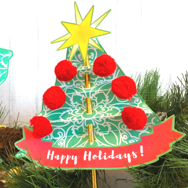 The paper Christmas tree straw craft made from Abbi Kirsten's free printables. The tree is made from green and white patterned paper. Red pompom adorn the sides of the tree and a a cut-out printed yellow straw is attached to the tree top. The Christmas tree craft says "Happy Holidays" on the bottom. 