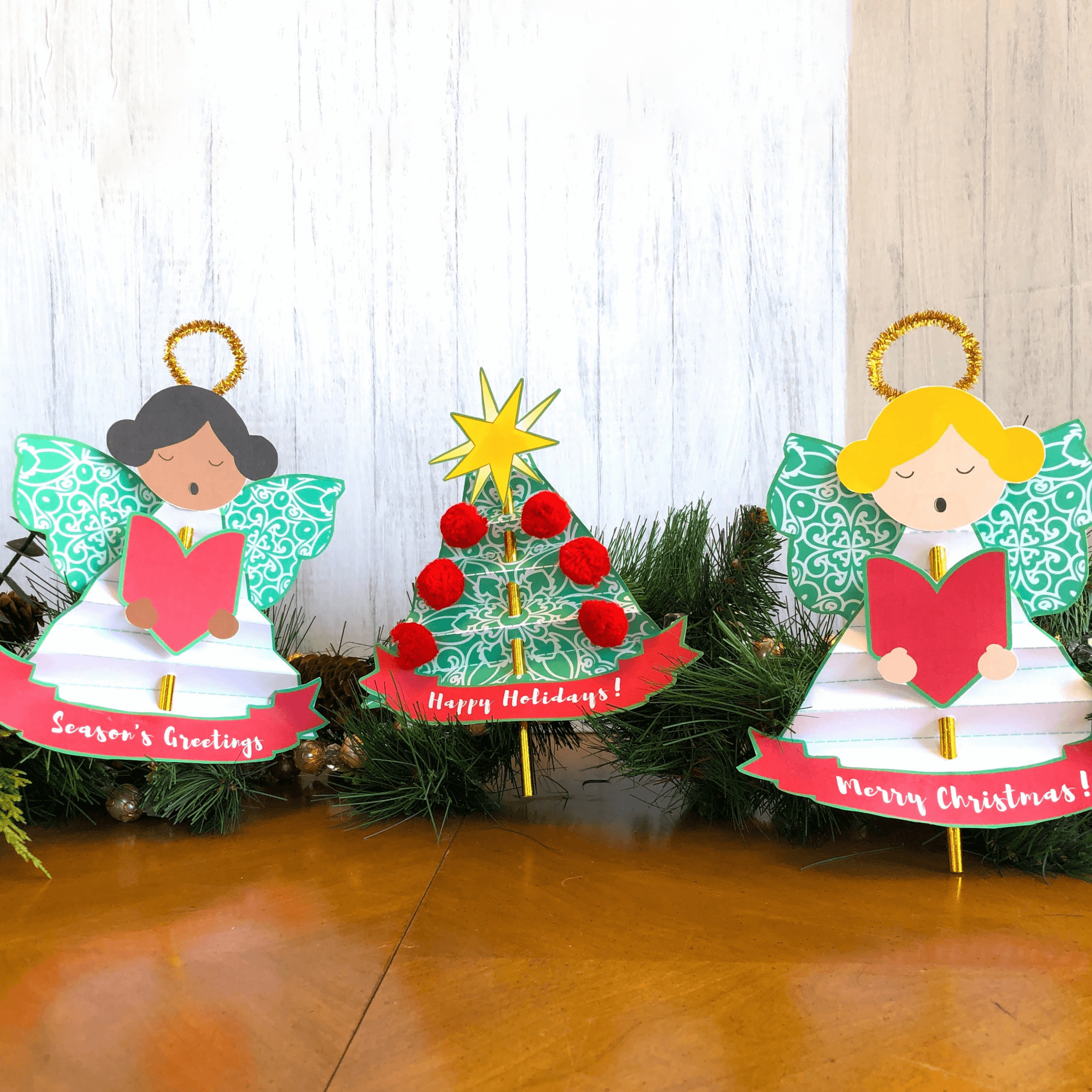 Two adorable singing angels and a Christmas tree made from folded paper and gold straws are lined up against a green pine garland. Banners at the bottom of the decorations read “Happy Holidays,” “Season’s Greetings,” and “Merry Christmas.”