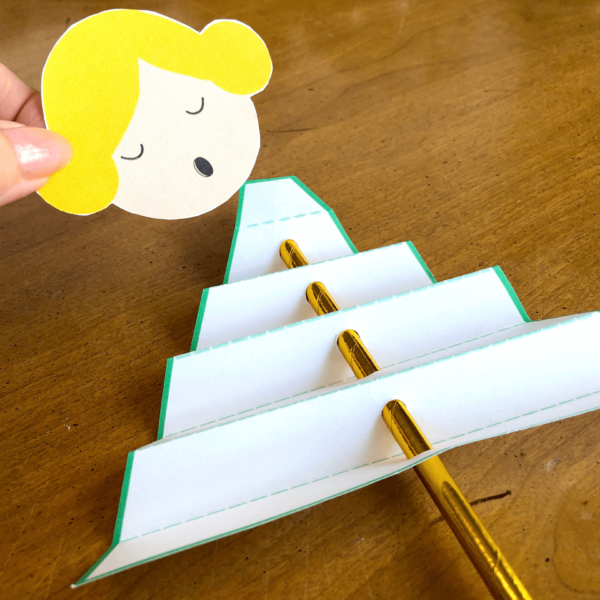 The paper Christmas angel craft is coming together. On the wooden table lays a white paper folded triangle with a gold paper straw threaded through it. Anni's hand is holding the blonde angel's head which needs to be glued to the top of the triangle. 