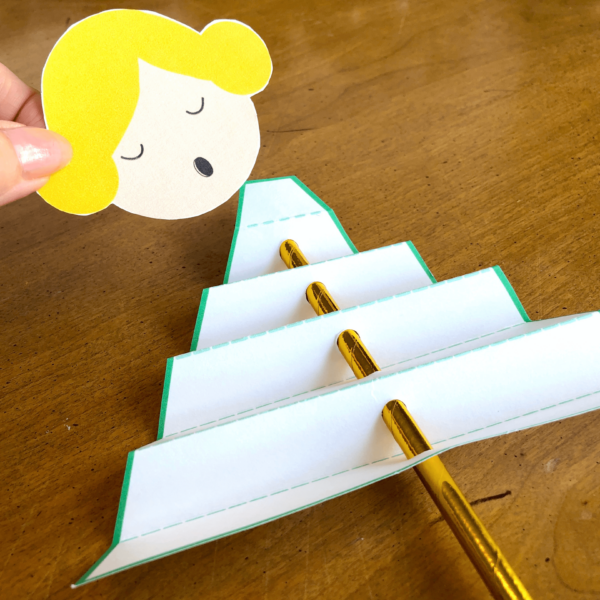 A person’s hand gently places the blonde angel head onto the triangle-shaped, accordion-folded paper body. 