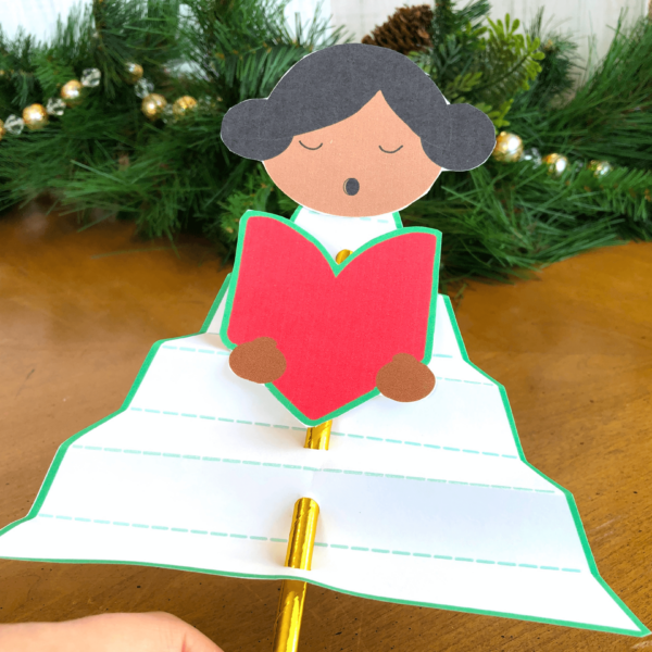 The black-haired paper Christmas angel craft from the front. Her hands and a red chorus book has been cut out and added to the front of the gold straw. 