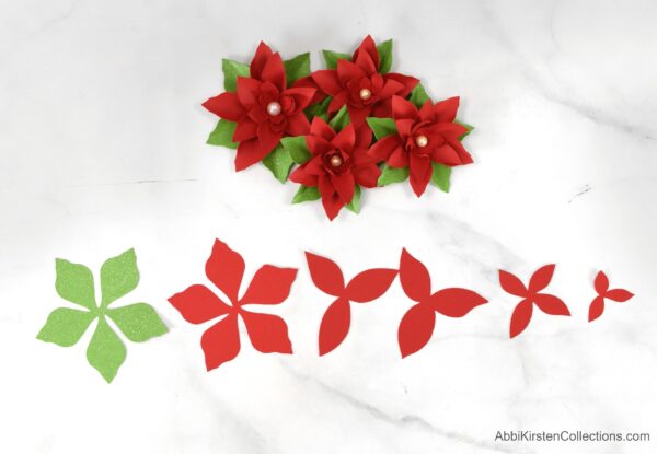 A green leaf base and poinsettia pieces lay in a line on a marble table below finished paper poinsettias.