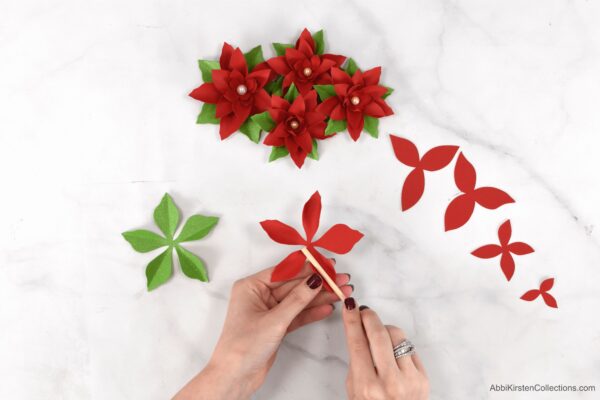Abbi Kirsten's hands roll the petals on the five-pointed poinsettia petal base. Additional pieces of the paper poinsettia craft and finished examples are also on the table.