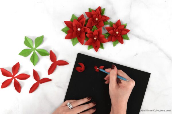 Abbi Kirsten's hands demonstrate how to shape poinsettia paper flowers using her Paper Blooms Shaping Mat and Toolset. Finished and unfinished paper poinsettias lay around the table. 