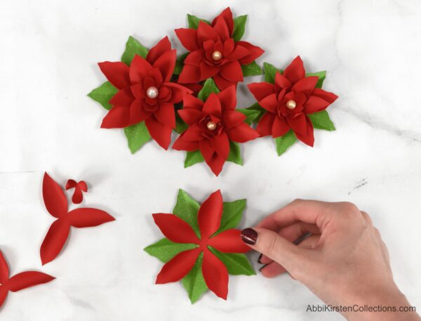 Abbi Kirsten's manicured hand places a piece of the red poinsettia onto the green leaves. Finished paper poinsettias and parts for the next step lay around the table.