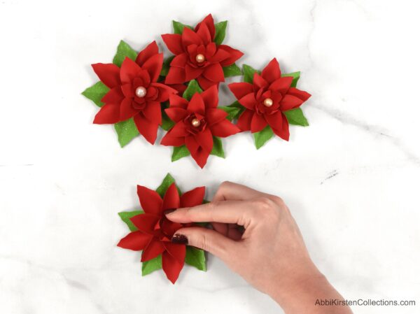 Four paper poinsettia flowers are gathered at the top of the photo, while Abbi Kirsten's hand places the small center leaves into the center of the paper poinsettia. 