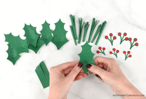 An overhead close-up of Abbi Kirsten’s hands holding a paper holly leaf. Additional pieces to make holly for a Christmas wreath lay in order on the table.