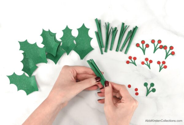 Abbi Kirsten rolls green paper to mimic pine needles for her DIY Christmas Wreath. Paper berries and holly leaves are also on the table, ready to be put together.
