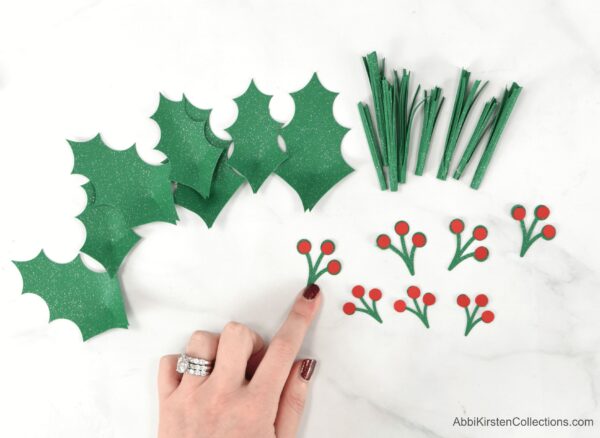 Abbi Kirsten's finger points to a paper berry piece of the DIY Christmas wreath. Holly leaves, paper pine needles and more red paper berries lay on the white table top.