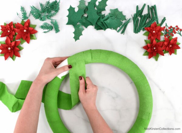 Abbi Kirsten wraps green crepe paper around a foam circle. Other Christmas wreath embellishments like paper pine needles, holly, and poinsettias lay nearby. 