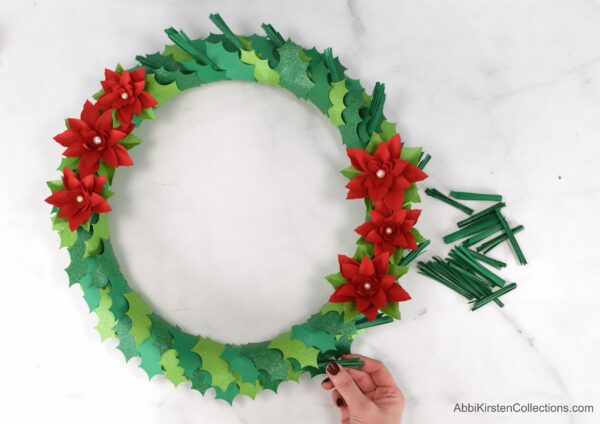 As part of the tutorial, Abbi shows how to glue paper pine needles onto a Christmas wreath covered in paper holly leaves and handmade poinsettia flowers.