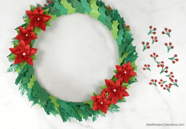 A handmade Christmas wreath is seen from above. Paper poinsettias are fixed on the wreath, and paper berries are on the right side of the wreath.