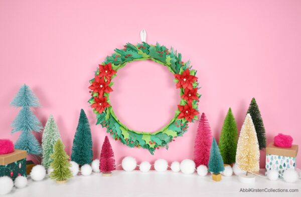 In front of a pink wall, on a white table, sit colorful Christmas trees, a string of white baubles, and small wrapped presents. A paper poinsettia and holly wreath hangs on the wall.