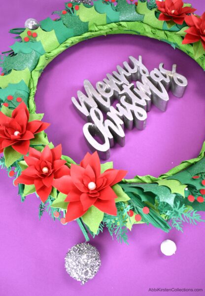 A close-up of a Christmas wreath with poinsettia paper flowers and holly lays on purple paper, with some silver ornament nearby and a sign in the middle of the  wreath that says "Merry Christmas." Use your Cricut machine to make a custom DIY holiday wreath out of paper.
