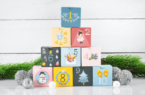 A stack of DIY advent calendar boxes to open on each day of December, surrounded by white baubles and garland. Download the free printable Christmas advent calendar boxes