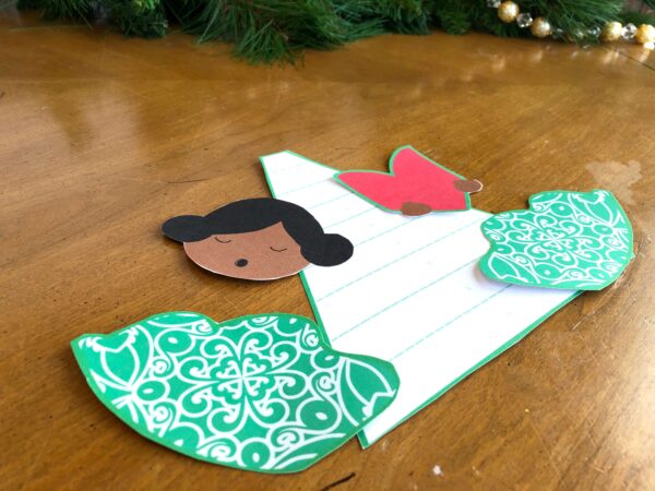 Separate cut-out pieces of a paper angel craft template are on a brown wooden table, including the cone-shaped angel body, green and white wings, a red songbook, and a small head with black hair.