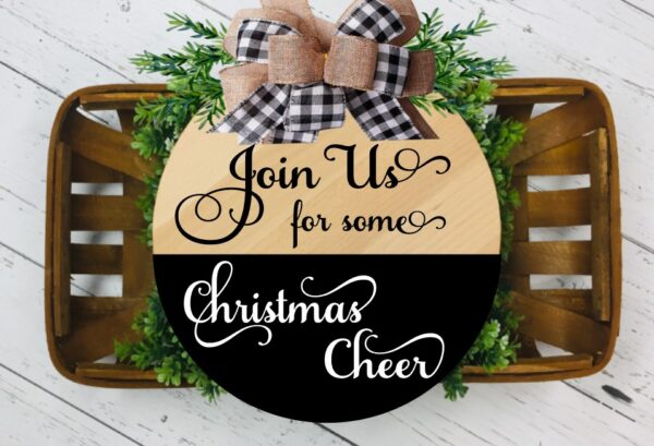 A tan, black and white Christmas Wood Door Hanger decorated with green foliage and a tan, black and white bow. 
