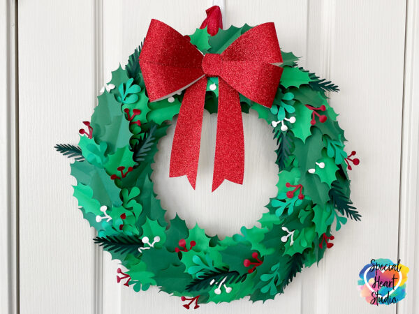 A green, red and white paper Christmas holly wreath decorated with a sparkly red bow hanging on a white door. 