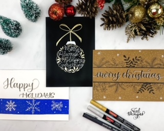 Three hand lettered holiday cards in a variety of styles on a white table next to Christmas decorations and three pens. 