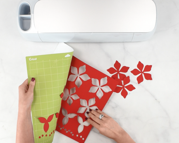 The top of a Cricut machine is next to Abbi Kirsten's hands as she rolls a green Cricut mat off red cardstock and poinsettia petals. 
