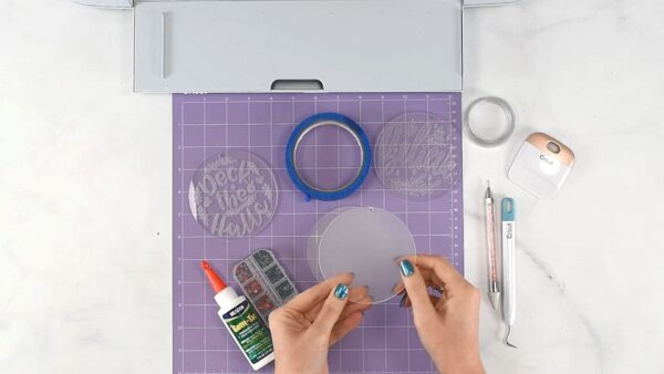 The supplies needed to make engraved acrylic Christmas ornaments are laid out on a purple cutting mat. You'll need acrylic disks, Cricut engraving tools, glue, some gems for decoration, and a Cricut cutting machine.