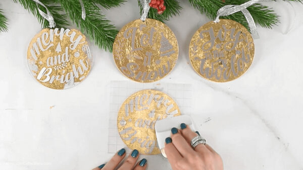 Abbi uses the Cricut scraper to thoroughly apply the vinyl decal to the front of a gold-gilded acrylic Christmas ornaments.