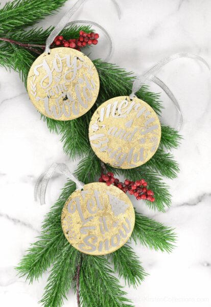 A set of three golf leaf acrylic ornaments, made with gilded gold leaf, white vinyl, and silver ribbons. The ornaments sit on faux evergreen branches with red holly berries.